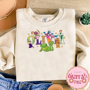 Könnte beinhalten: Cremefarbenes Sweatshirt mit dem Namen "OLIVIA" in bunten, cartoonartigen Buchstaben und Muppet-Charakteren. Das Design umfasst Kermit den Frosch, Fozzie Bär und andere Muppet-Charaktere. Das Sweatshirt hat einen Rundhalsausschnitt.