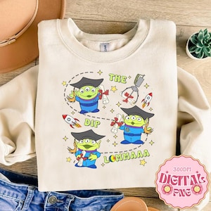 Op de afbeelding: Crèmekleurige sweatshirt met een afbeelding van groene aliens met afstudeerhoeden, diploma's vasthoudend en omringd door sterren en raketten. De tekst bevat "THE DIP LOMMAAA". Er is ook een grijpmachine afgebeeld.