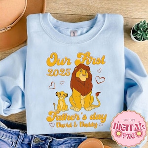 Puede incluir: Sudadera azul claro con un gráfico de un león y un cachorro, con el texto "Our First 2025 Father's Day David & Daddy". El diseño es en amarillo y naranja. Un gráfico PNG digital está en la esquina inferior derecha.