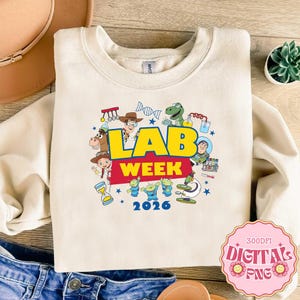 Könnte beinhalten: Cremefarbenes Sweatshirt mit einem farbenfrohen Aufdruck „LAB WEEK 2026“. Das Design zeigt Zeichentrickfiguren und Illustrationen zum Thema Wissenschaft. Das Sweatshirt hat einen Rundhalsausschnitt.