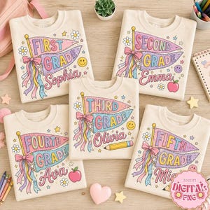 May include: Five cream-colored sweatshirts featuring colorful pennant flag graphics with text reading First, Second, Third, Fourth, and Fifth Grade, along with names Sophia, Emma, Olivia, Ava, and Mia, decorated with flowers, stars, and pencils.