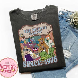 Może przedstawiać: Ciemnoszara koszulka z grafiką postaci z Muppet Show z napisem "The Muppet Show Since 1976". Koszulka jest prezentowana na niebieskich szortach dżinsowych.