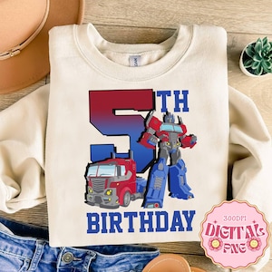 May include: Cream-colored sweatshirt featuring a red and blue graphic of Optimus Prime and a truck. The number "5TH" is displayed above the graphic, with the word "BIRTHDAY" below. A pink sticker with "DIGITAL PNG" is in the bottom right corner.