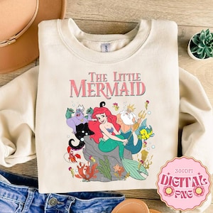 May include: Cream-colored sweatshirt featuring a graphic of Ariel, Ursula, King Triton, and Flounder from The Little Mermaid. The words "The Little Mermaid" are printed in pink. The design includes coral, seaweed, and bubbles.