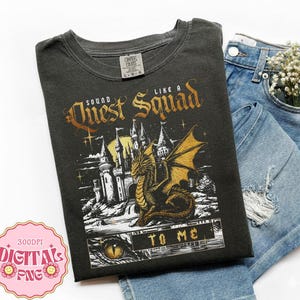 Op de afbeelding: Een antracietgrijs t-shirt met een grafische print van een kasteel en een draak met de tekst "SOUND LIKE A QUEST SQUAD TO ME". Het t-shirt wordt gecombineerd met een lichtblauwe, versleten spijkerbroek en witte bloemen.