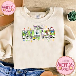May include: Cream-colored sweatshirt featuring the name "HUNTER" in colorful, cartoon-style letters. The letters are decorated with images of Buzz Lightyear and other Toy Story characters, with stars and a rocket ship. The sweatshirt has a crew neck.