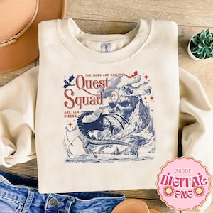 Op de afbeelding: Een crèmekleurige sweater met ronde hals en een grafische print van een draak die over een bergketen met een schedelvorm vliegt. De tekst luidt "THE ISLES ARE CALLING QUEST SQUAD ARETIAN RIDERS" in rode en blauwe inkt met steraccenten.