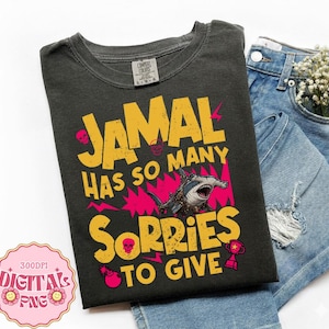 May include: A charcoal gray t-shirt featuring the yellow text JAMAL HAS SO MANY SORRIES TO GIVE, accented with pink lightning bolts, small skulls, a hammerhead shark illustration, a trophy, and a bomb icon, paired with blue denim jeans.