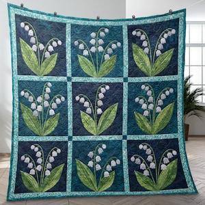 以下が含まれることがあります： スズランの花の繰り返し模様が特徴のキルトブランケット。各正方形には、白いベル型の花と緑の葉が、濃いティール色の背景に配置され、ターコイズのボーダーで縁取られています。キルトは壁に飾られています。