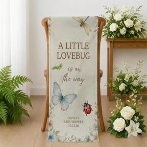 May include: A cream-colored baby shower banner draped over a wooden chair. The banner features a dragonfly, caterpillar, and butterfly illustrations, along with the text "A Little Lovebug is on the Way." The banner is decorated with floral accents and the text "Olivia's Baby Shower 10.12.26."