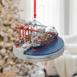 May include: A Christmas ornament depicting a miniature ocean freight dock scene. The ornament features a cargo ship, cranes, containers, and a dock. The text "Robert's Ocean Freight Dock - 2025" is printed on the base. A red ribbon is attached for hanging.
