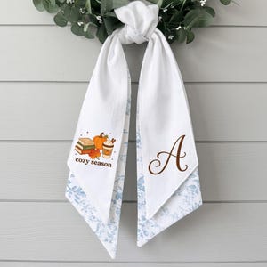 May include: A white fabric scarf with a blue floral pattern on the reverse side. One end features a stack of books, a pumpkin, and a coffee cup with the text "cozy season". The other end displays a large brown script letter "A" in the center.
