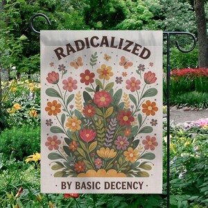 Op de afbeelding: Een beige tuinvaandel met de woorden "RADICALIZED" en "BY BASIC DECENCY" in bruin. De vlag heeft een bloemenontwerp met roze, oranje en gele bloemen, groene bladeren en vlinders.