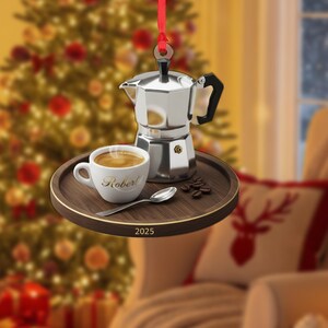 May include: A Christmas ornament featuring a silver coffee maker, a white coffee cup with the name "Robert", a spoon, and coffee beans on a wooden tray. The tray has the year "2025" printed on it. The ornament hangs from a red ribbon.