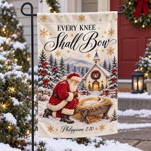 May include: A rectangular garden flag featuring a winter scene with Santa Claus kneeling before a manger containing a baby. The text reads Every Knee Shall Bow and Philippians 2:10. Gold snowflakes decorate the cream-colored background.
