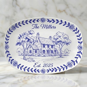 Puede incluir: Plato ovalado blanco con borde floral azul e ilustración azul de una casa y árboles. El texto "The Millers" y "Est. 2025" también es azul.