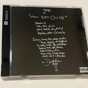 May include: A black CD case for the 2Pac album "All Eyez On Me." The album title and artist name are in white text. The case also lists the producers and featured artists. The case is labeled "2 DISCS."