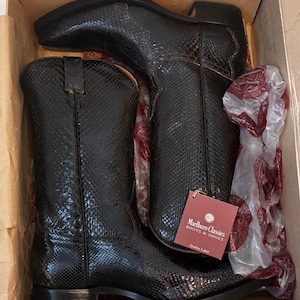 Op de afbeelding: Een paar zwarte cowboylaarzen met een slangenpatroon, gepresenteerd in een kartonnen doos. De laarzen hebben een puntige neus en een gestapelde hak. Een label van "Marlboro Classics Boots & Shoes" is bevestigd.