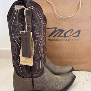 May include: Dark brown leather cowboy boots featuring a textured, speckled metallic gold vamp and shaft accents. A brown cardboard tag attached with twine displays the text Marlboro Classics Boots & Shoes. The boots have decorative white stitching.