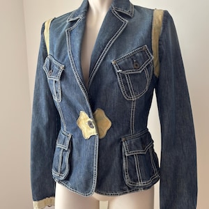 Pode incluir: Um blazer de ganga azul escuro com painéis de camurça bege claro nos ombros, punhos e fecho frontal. O casaco inclui quatro bolsos com aba e botões, costuras brancas em contraste e um fecho frontal com um único botão.