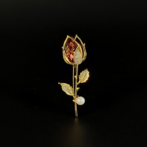 May include: A gold-toned rose brooch featuring a large, faceted, reddish-pink crystal bud encased in gold petals encrusted with small clear stones. The stem includes two gold leaves covered in clear stones and a single white spherical pearl accent.