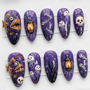 May include: A set of ten purple press-on nails with Halloween-themed designs. The designs include a fish, a lantern, a skull, jellyfish, and fish bones. The nails are in a clear plastic case.