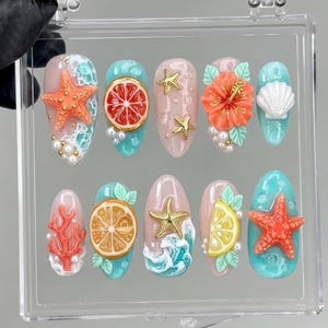 May include: A set of ten press-on nails featuring a tropical theme with 3D embellishments including orange and lemon slices, starfish, coral, hibiscus flowers, seashells, and white pearls on a mix of light pink and turquoise blue backgrounds.