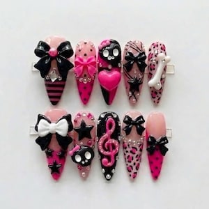 May include: A set of ten press-on nails featuring a pink and black color palette. Designs include 3D black and white bows, pink hearts, black skulls, stars, a pink musical treble clef, leopard print patterns, and small clear rhinestones.