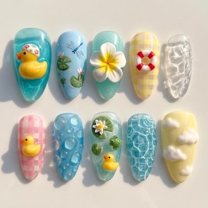 May include: A collection of ten artificial nails with various summer-themed designs. The designs include a yellow rubber duck, a dragonfly, a white flower, a life preserver, water droplets, a pink gingham pattern, bubbles, a frog, and clouds.