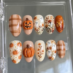 Può includere: Un set di dieci unghie finte con motivi a tema autunnale, tra cui fantasie a quadri marroni, zucche arancioni in rilievo 3D, fantasmi bianchi, motivi floreali arancioni e marroni e foglie, disposte in un contenitore di plastica trasparente.