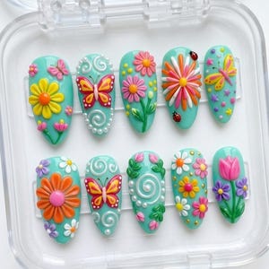 May include: A set of ten almond-shaped press-on nails with a light teal base, featuring 3D raised designs including colorful butterflies, various flowers like daisies and tulips, white swirls, small ladybugs, and tiny yellow stars.
