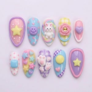 May include: A collection of ten colorful, decorated artificial nails. Designs include a yellow star, a smiling face, a white bunny, a pink flower, a purple heart, a blue square, stars, hearts, paw prints, and a yellow circle.