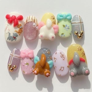 Può includere: Un set di dieci unghie finte decorate con vari disegni: fiocchi, animali dei cartoni animati, glitter e note musicali. I colori includono bianco, rosa, giallo e grigio. Alcune unghie presentano elementi 3D.