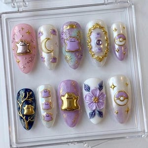 May include: A set of ten almond-shaped press-on nails featuring purple blob characters, gold metallic accents, celestial motifs, a gold ornate frame, a purple flower, and spherical icons in a clear plastic display case with a white background.