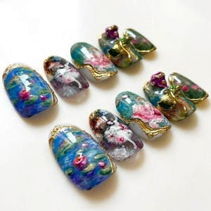 Puede incluir: Una colecci&oacute;n de u&ntilde;as artificiales con dise&ntilde;os art&iacute;sticos. Las u&ntilde;as presentan pinturas en miniatura de nen&uacute;fares, arreglos florales y detalles dorados ornamentados. La paleta de colores incluye azules, verdes, rosas y morados, creando una est&eacute;tica delicada y elegante.