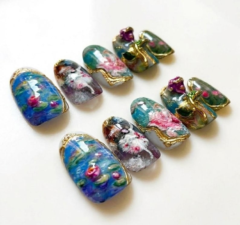 Puede incluir: Una colecci&oacute;n de u&ntilde;as artificiales con dise&ntilde;os art&iacute;sticos. Las u&ntilde;as presentan pinturas en miniatura de nen&uacute;fares, arreglos florales y detalles dorados ornamentados. La paleta de colores incluye azules, verdes, rosas y morados, creando una est&eacute;tica delicada y elegante.