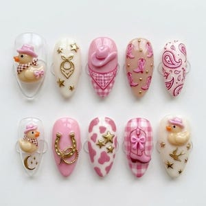 May include: A set of ten acrylic nails with a western theme. Designs include a duck with a cowboy hat, horseshoes, cow print, cowboy boots, and paisley patterns. Colors are pink, white, and gold.