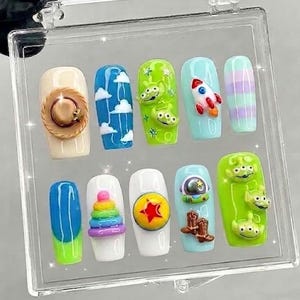 Könnte beinhalten: Ein Set aus zehn künstlichen Nägeln mit verschiedenen Designs. Die Designs umfassen einen Cowboyhut, Wolken, grüne Aliens, eine Rakete, Streifen, eine blau-grüne Spirale, ein Regenbogen-Eis, einen gelb-roten Ball, einen Weltraumhelm und Cowboy-Stiefel.