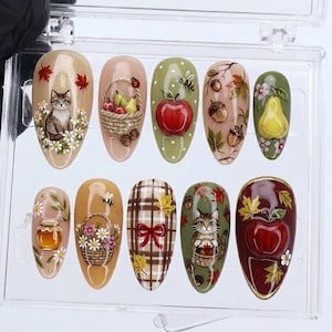 May include: A set of ten press-on nails featuring autumn-themed designs including a cat, a basket of fruit, a red apple, acorns, a pear, a honey jar, a flower basket, a plaid pattern with a red bow, and fall leaves in a clear plastic case.