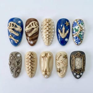May include: A set of ten artificial nails with a dinosaur theme. Designs include dinosaur skeletons, footprints, and bone structures in various colors like blue, brown, and beige. Some nails feature pearl accents and gold stars.