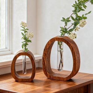 Op de afbeelding: Twee houten vazen met een cirkelvormig ontwerp, elk met een glazen buis voor het presenteren van bloemen. De vazen zijn gemaakt van donker hout en staan op een houten tafel. Eén vaas heeft een kleine tak met witte bloemen, de andere een langere tak.