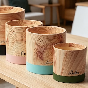 May include: Four wooden cylindrical planters with a natural wood grain finish. Each planter has a colored base: black, pink, teal, and green. The planters are engraved with names: Aria, Co, Luca, and Bella. They are arranged on a light wood table.