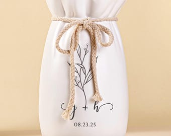 May include: A white ceramic vase with a drawstring design, tied with a beige rope. The vase features a black floral design and the date 08.23.25. The vase is filled with dried stems and sits on a woven coaster.