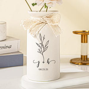 Puede incluir: Un jarrón de cerámica blanco con un diseño floral y un lazo de encaje, que contiene una flor rosa claro y vegetación. El jarrón tiene las iniciales "j + h" y la fecha "08.23.25" impresas. Una vela y libros están en el fondo.