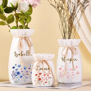 May include: Three white ceramic vases with floral designs and the names "Isabella", "Emma", and "Harper" in gold script. Each vase has a beige rope tie around the neck. The vases are filled with flowers and dried plants.