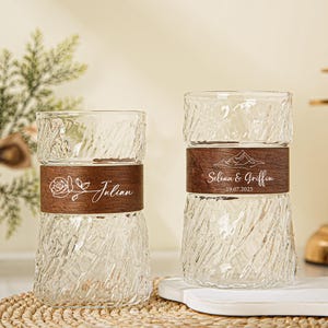 Op de afbeelding: Twee heldere glazen bekers met een textuurontwerp en houten banden. Eén beker heeft een bloemenontwerp en de naam "Julian", de andere heeft een bergontwerp en de namen "Selina & Griffin" en de datum "19.07.2025".