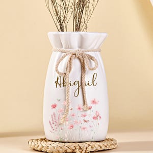 Puede incluir: Jarrón de cerámica blanca con diseño floral y el nombre "Abigail" en escritura cursiva. El jarrón está atado con un detalle de cuerda y se apoya en un posavasos tejido. Hay flores secas dentro.