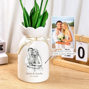 May include: A white ceramic vase with a scalloped rim, tied with a gold ribbon. The front features a black and white line drawing of a couple, the names James & Amelia, and the date 12.11.2026. A small inset photo shows the original couple.