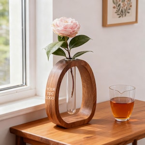 Pode incluir: Um vaso de madeira com um design oval único, segurando uma única flor rosa e folhas verdes. O vaso tem um tubo de vidro transparente para a flor e o texto "YOUR TEXT LOGO" está impresso na lateral. Um copo de chá está na mesa de madeira.