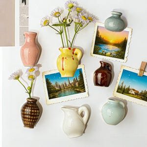 May include: A collection of miniature ceramic vases in various colors and styles, including pink, yellow, brown, and white. Some vases hold small white and yellow flowers. Vintage-style postcards are also displayed.